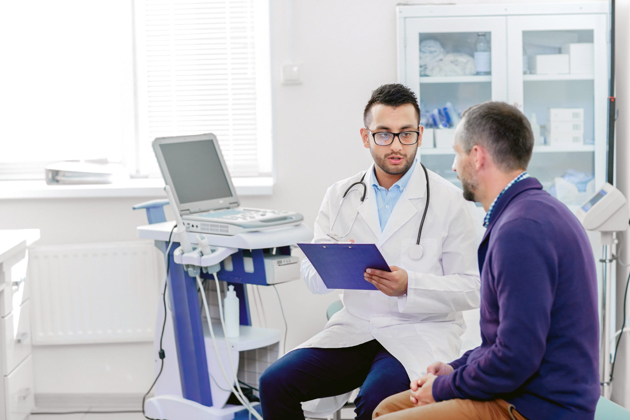 A light-skinned doctor in a white lab coat sitting with a light-skinned patient wearing a purple sweater. The doctor is consulting with the patient in a bright hospital room while holding a clipboard.