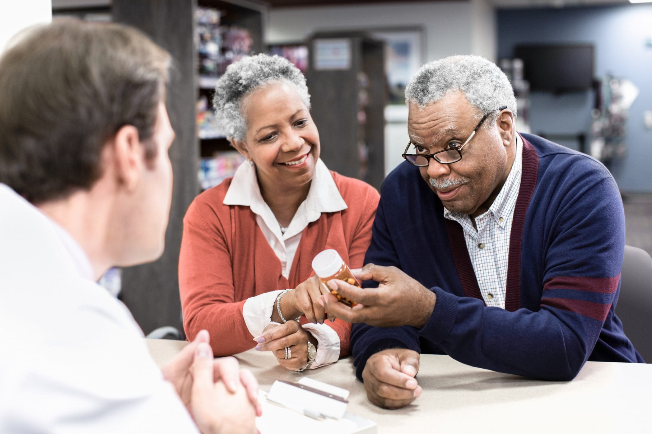 A light-skinned pharmacist consulting with two elderly, dark-skinned customers who are leaning over a counter with their elbows while reading a prescription bottle.