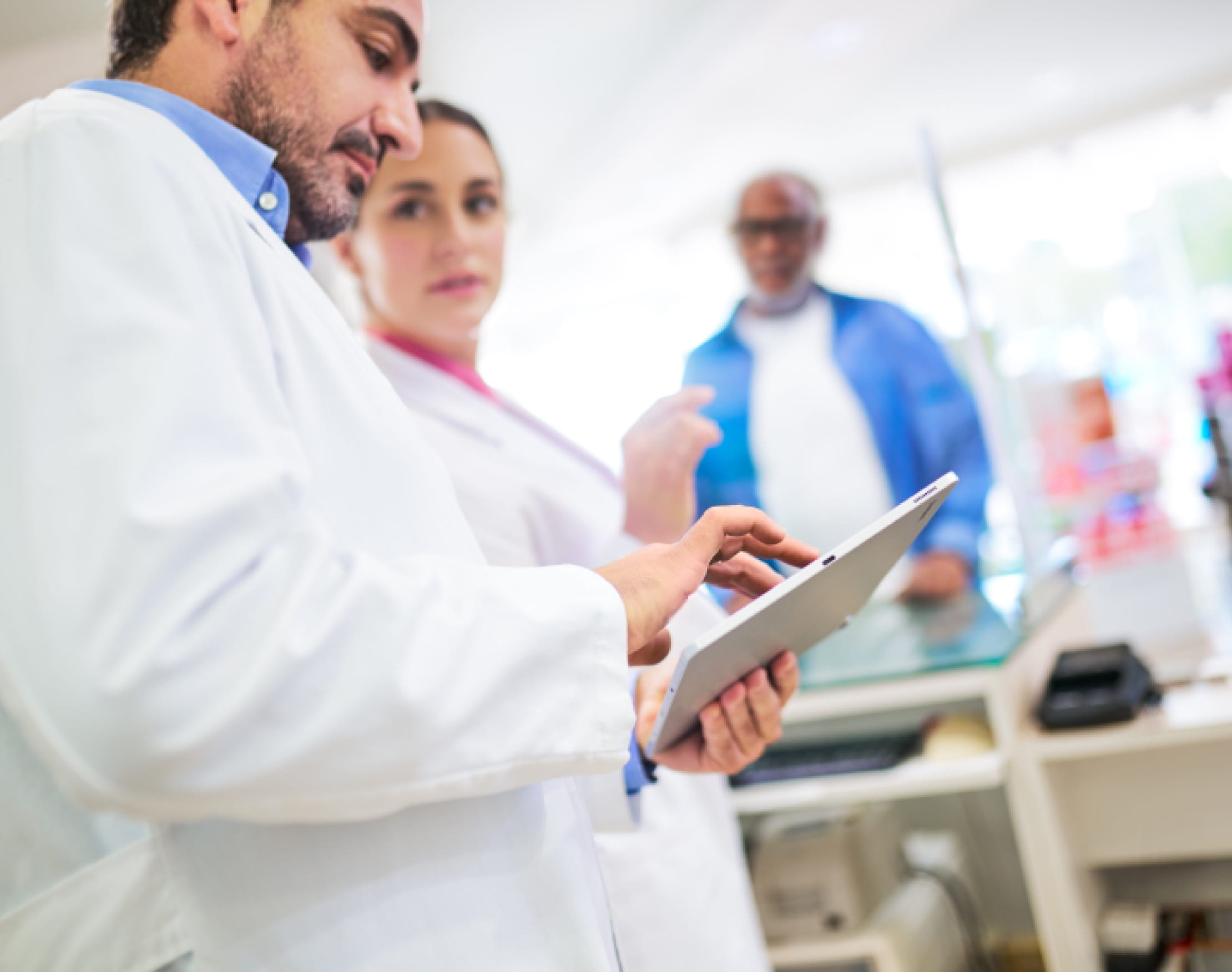 A pharmacist holding a tablet device consulting with another pharmacist while a patient waits behind the pharmacy counter.