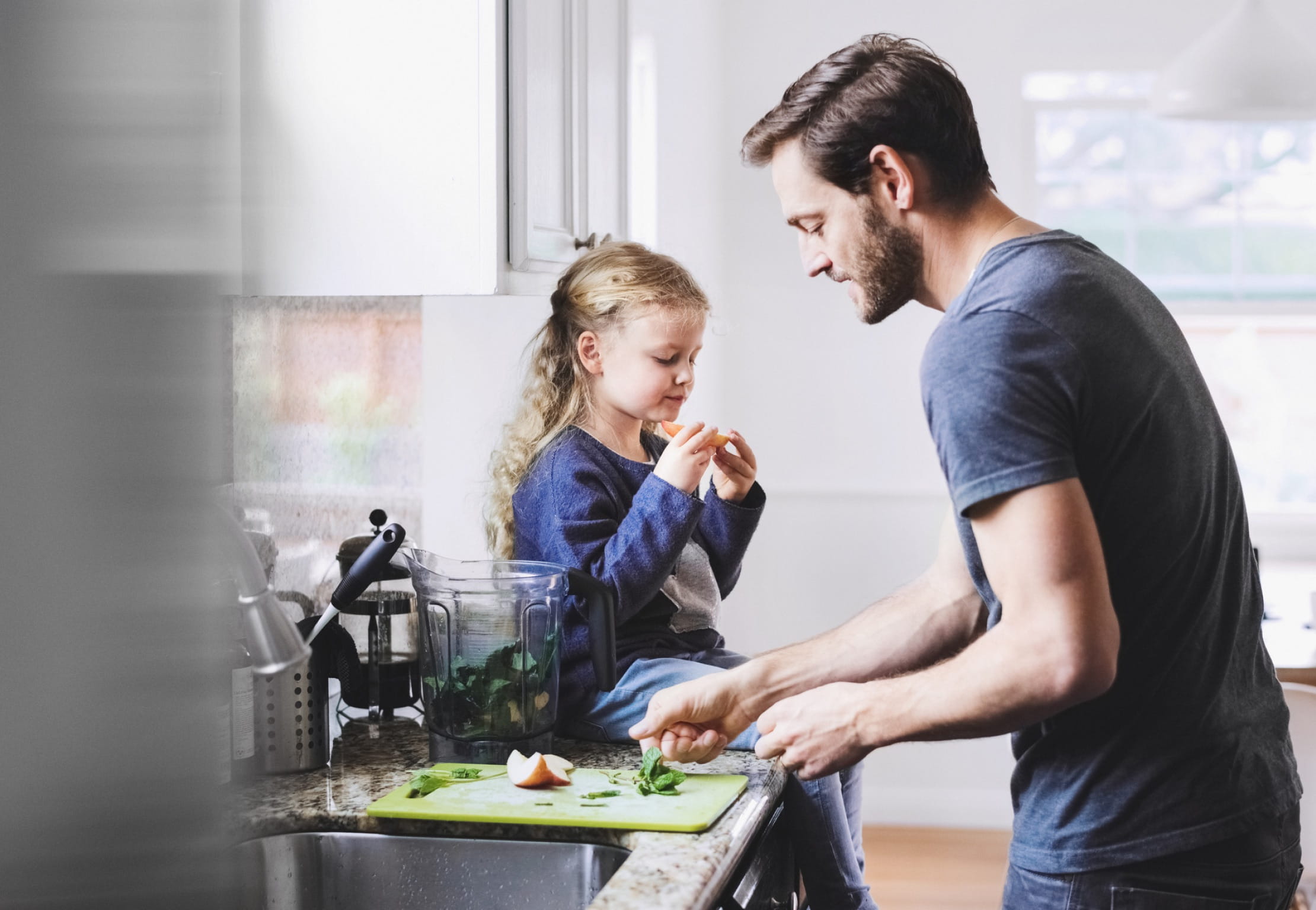 A parent preparing a snack on a kitchen counter next to their child who sits on the counter eating a slice of apple.