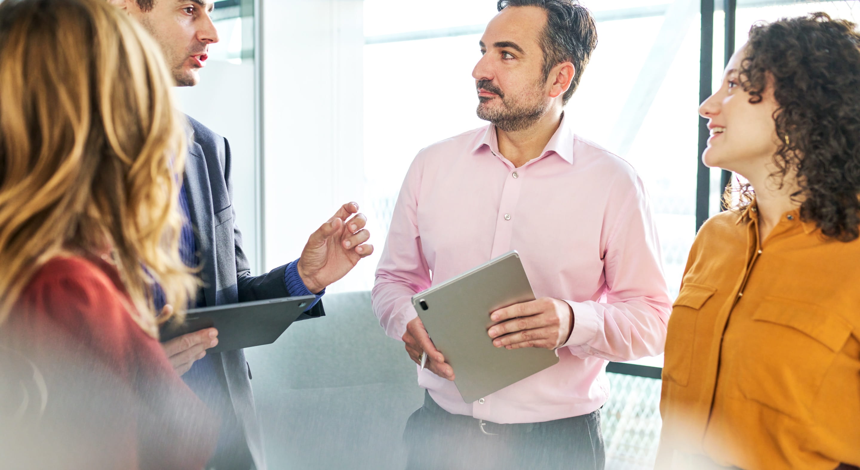 Four people standing and conversing in front of a bright window in an office. Two of them hold tablet devices in their hands.