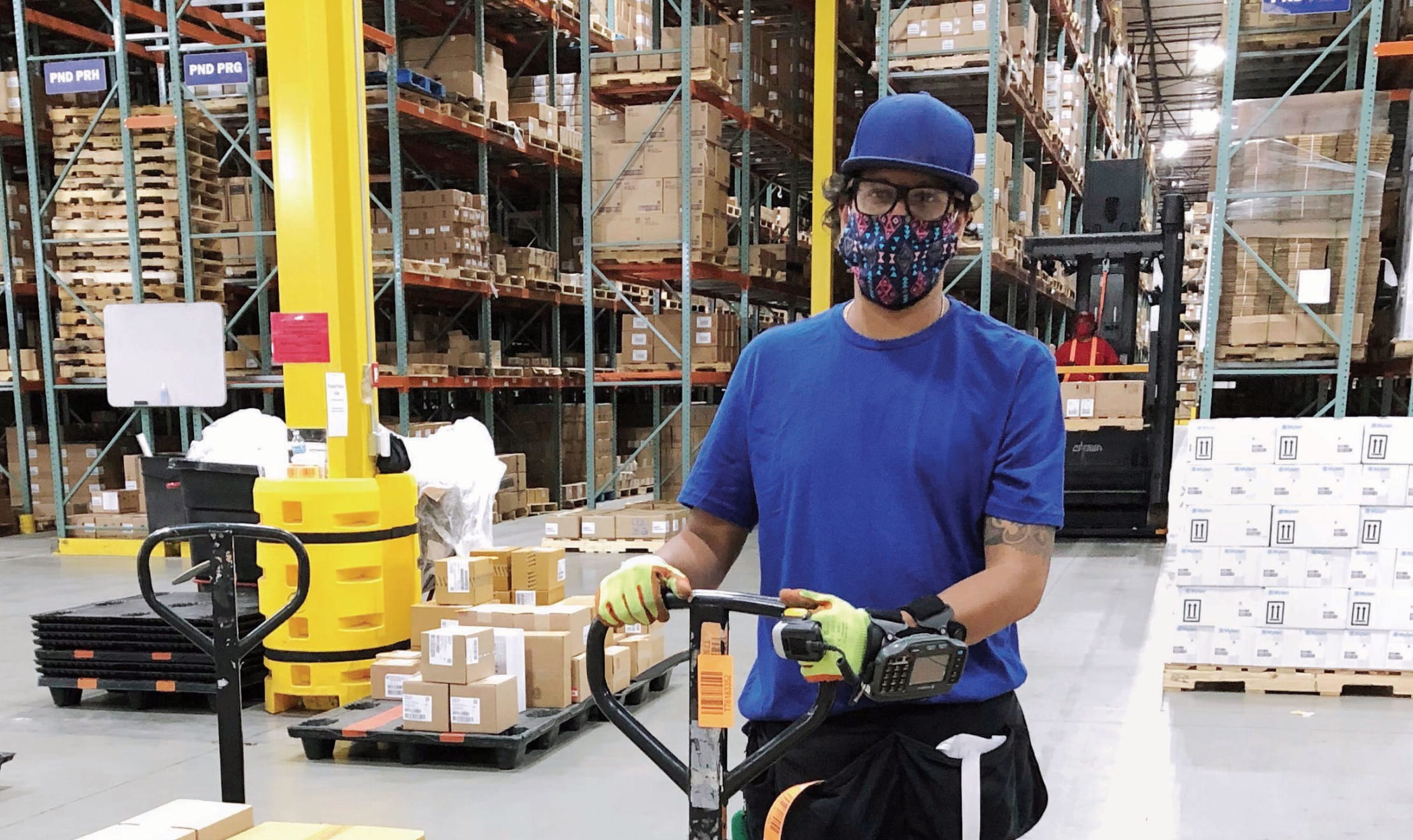 A warehouse worker operating a small forklift in a warehouse surrounded by boxes. They wear a blue shirt, blue hat, and a blue mask that covers their face and nose.