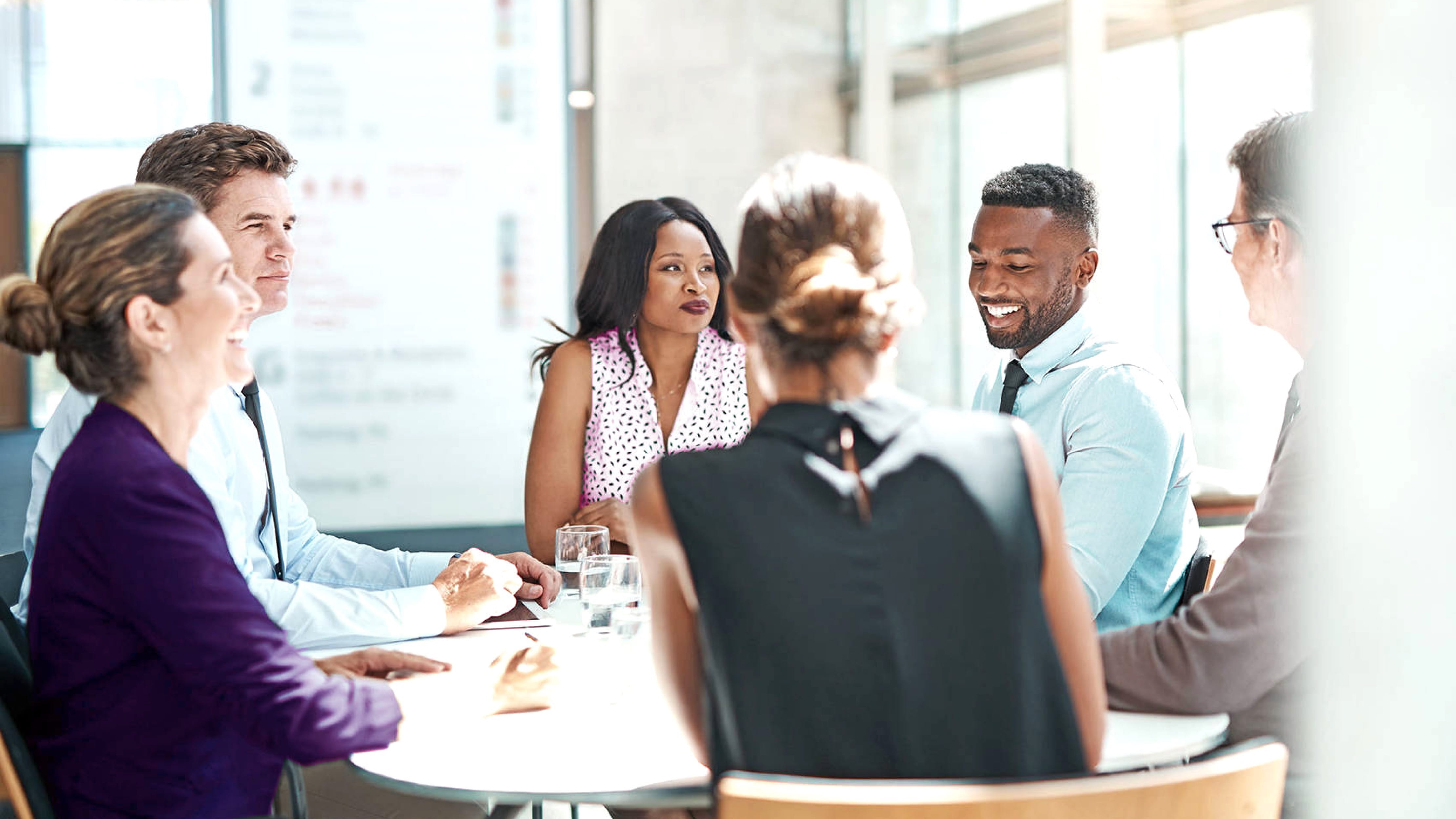 A diverse group of six office workers are gathered in a bright meeting room.