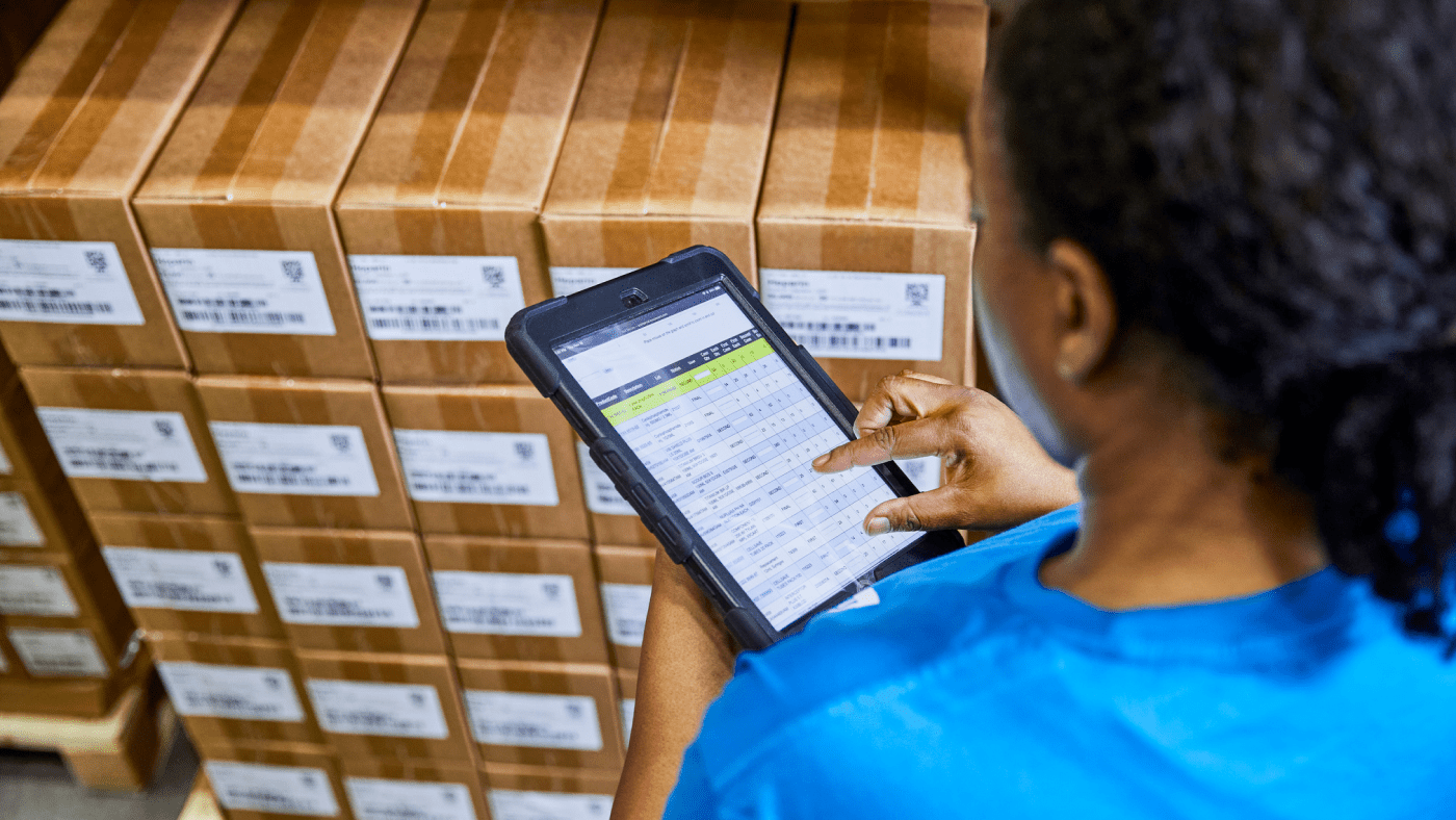 A person reviewing inventory data on a small tablet device. They wear a blue t-shirt and stand in front of a pallet of boxes that each have barcode labels attached.