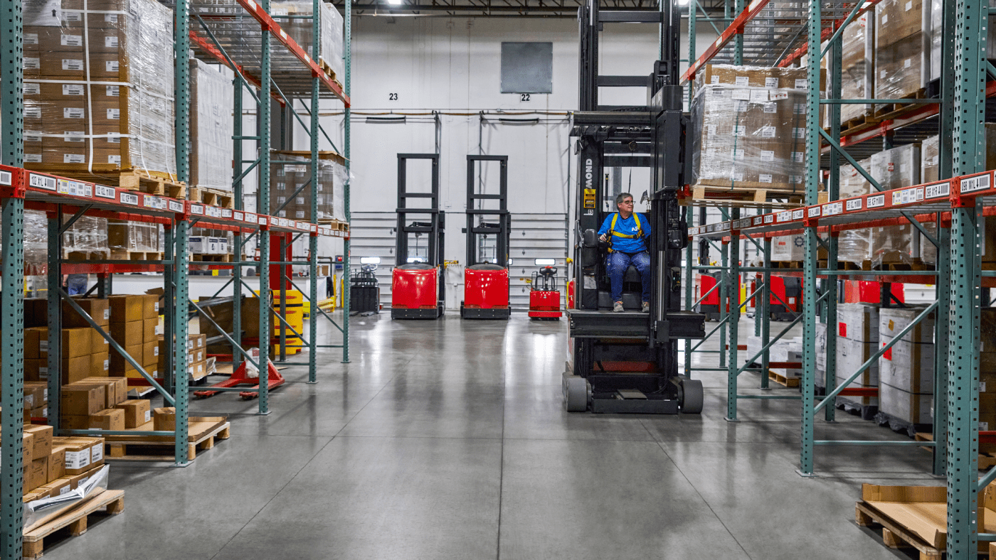 A worker wearing a blue shirt and yellow safety straps operating a forklift inside of a warehouse. They are using the equipment to place a pallet of boxes on an upper shelf.
