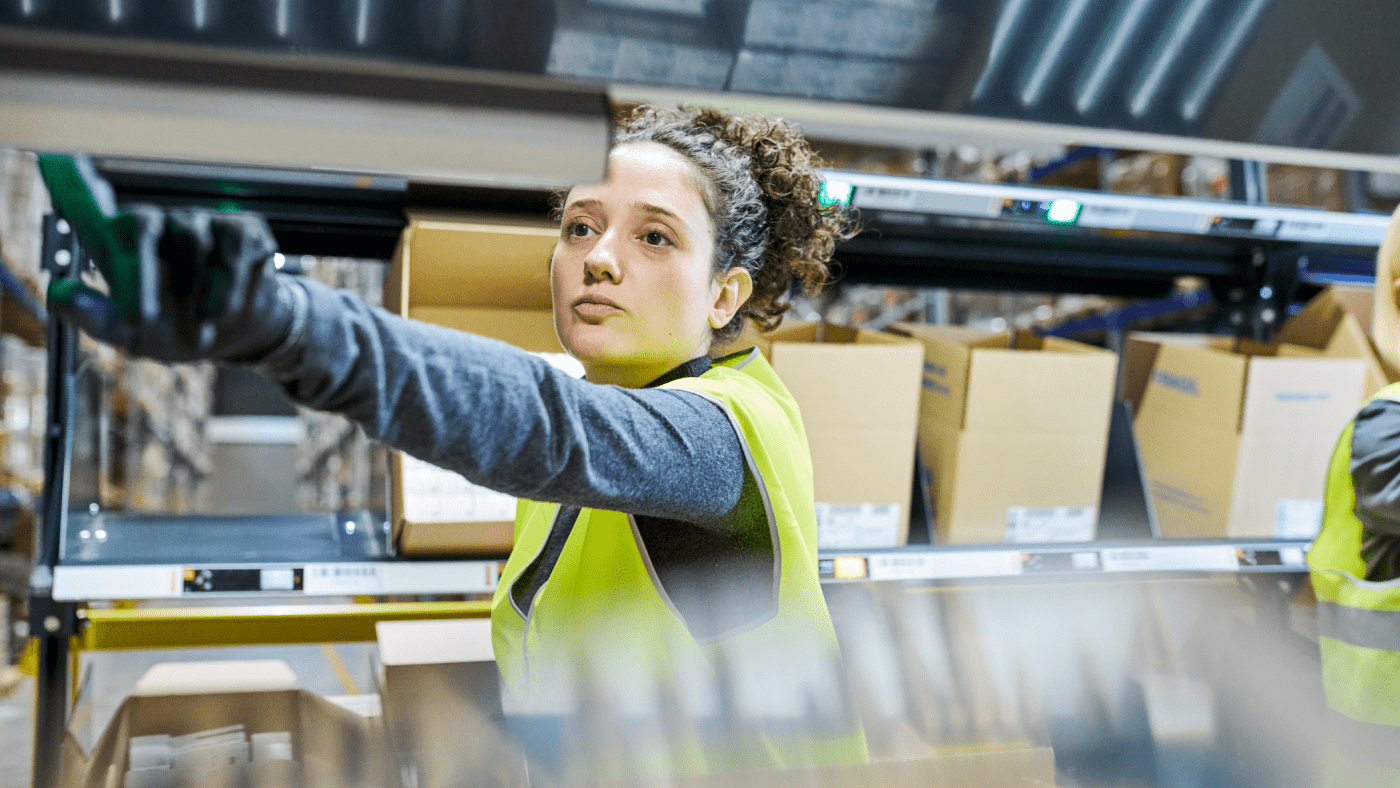 A worker pressing a button on a shelf inside a warehouse. They wear a yellow vest, a dark long-sleeve shirt, and gloves.