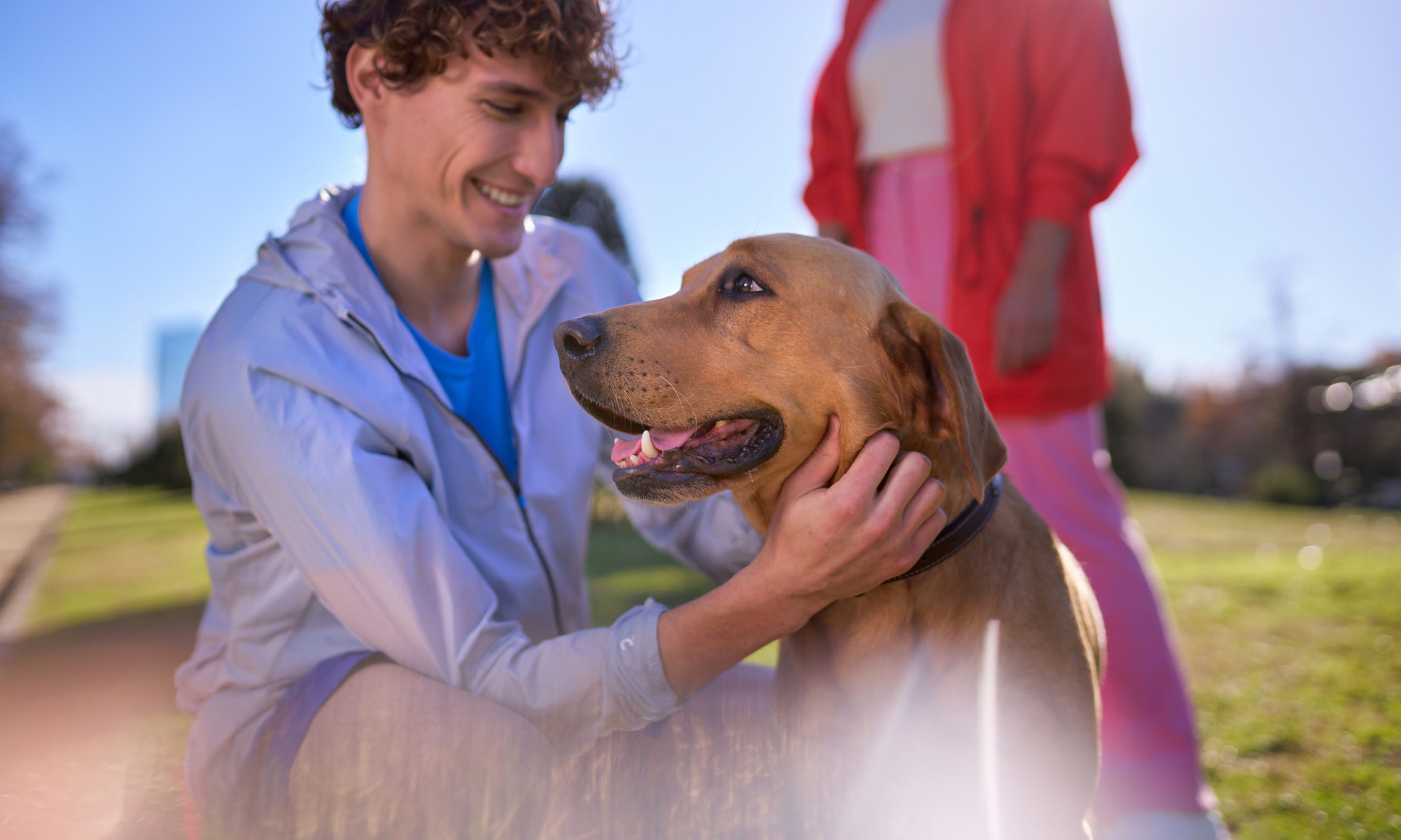 A light-skinned person in a white track jacket kneeling down to pet a dog in an outdoor park setting during a sunny day.