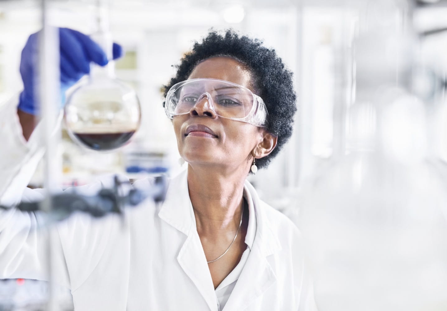 A dark-skinned lab technician in a white jacket and protective eye goggles looking intently at a beaker they are holding that is half full with dark liquid.