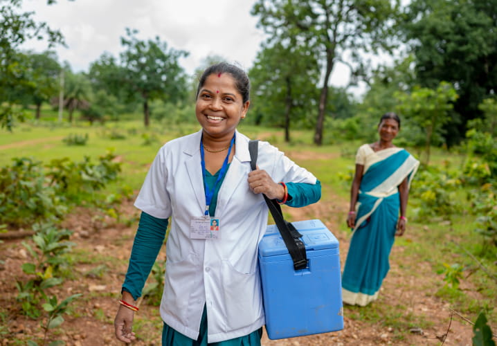 A dark-skinned medical professional smiling in an outdoor rural setting with lots of green vegetation holds a medical travel cooler. Behind them a dark-skinned person smiles and wears a green and white wrapped dress.