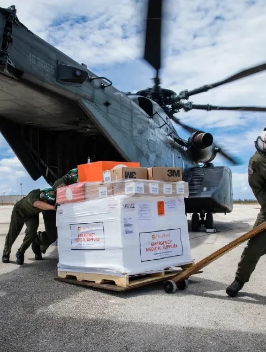 Workers in protective gear offload a pallet of supplies from a helicopter. Some of the boxes have 3M logos printed on them and others have text that reads, “Emergency Medical Supplies.