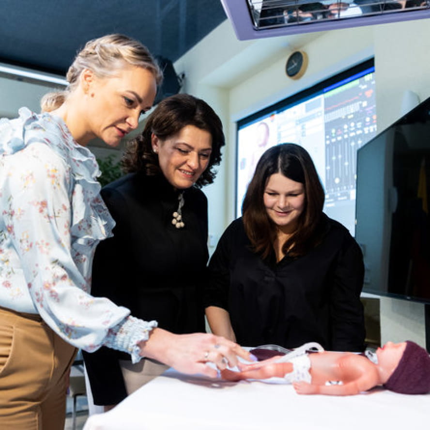 Three people looking down at a doll-like infant simulator laying on a table. The person on the left is pointing to a part of the device as the others watch.