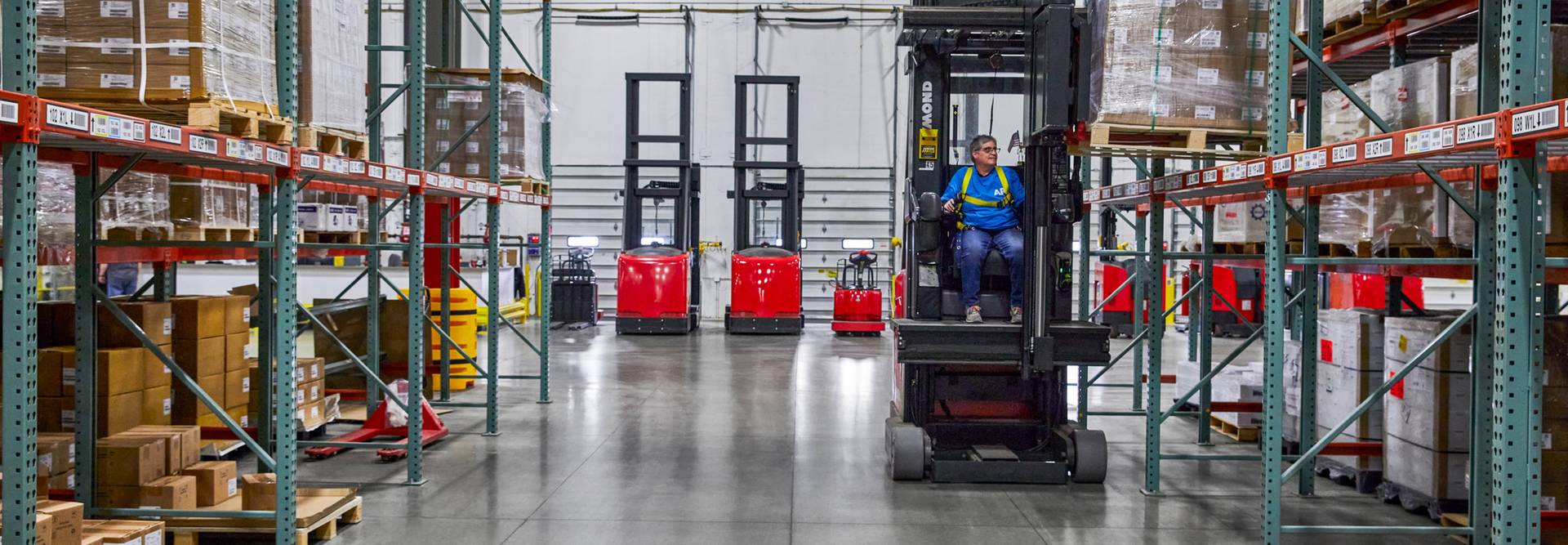 A worker wearing a blue shirt and yellow safety straps operating a forklift inside of a warehouse. They are using the equipment to place a pallet of boxes on an upper shelf.