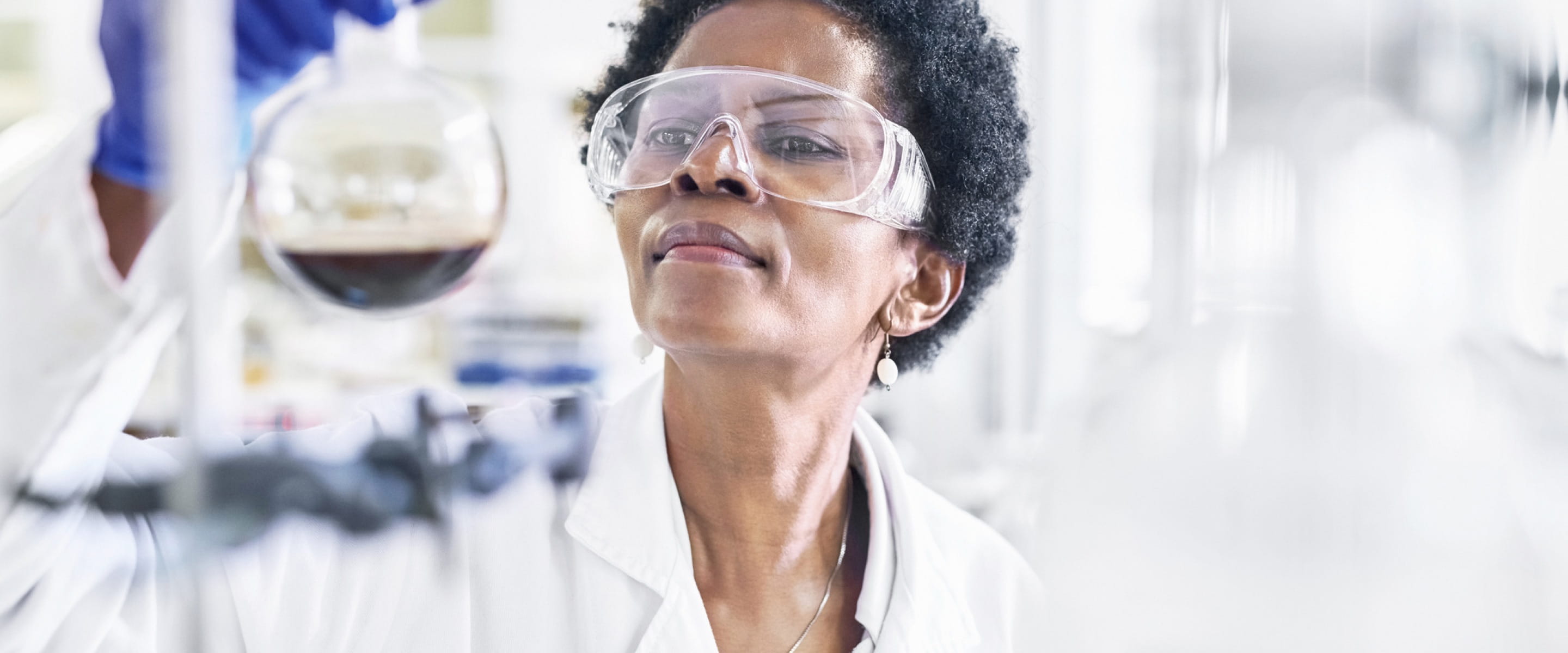 A dark-skinned lab technician in a white jacket and protective eye goggles looking intently at a beaker they are holding that is half full with dark liquid.