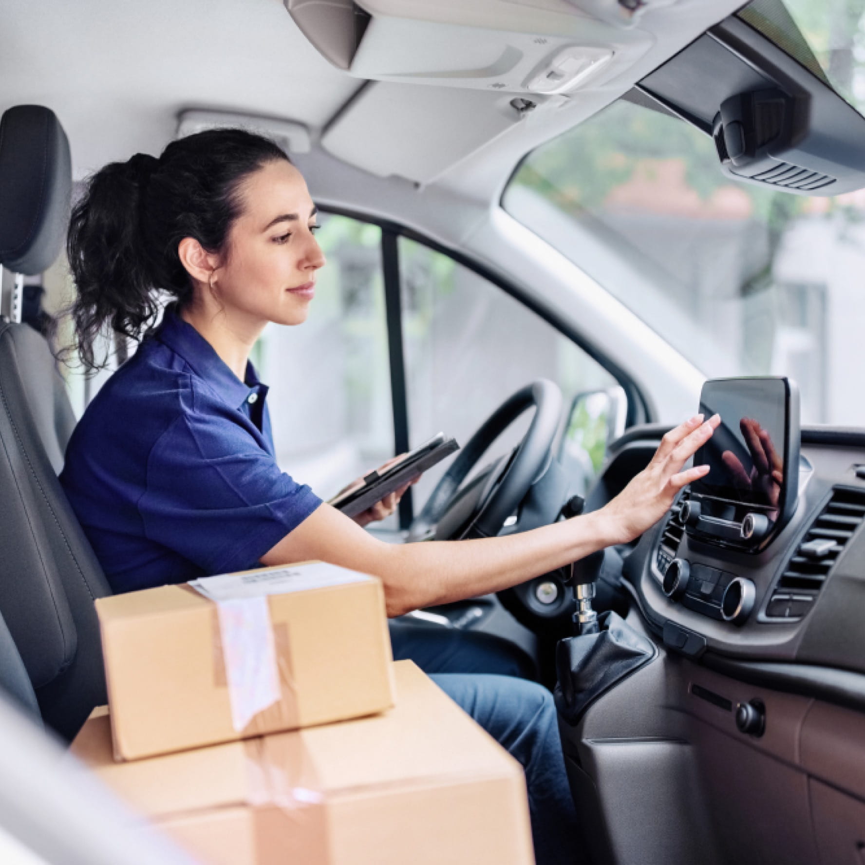 A delivery driver typing an address into the dashboard screen of their delivery van. Cardboard boxes are stacked up on the passenger seat.