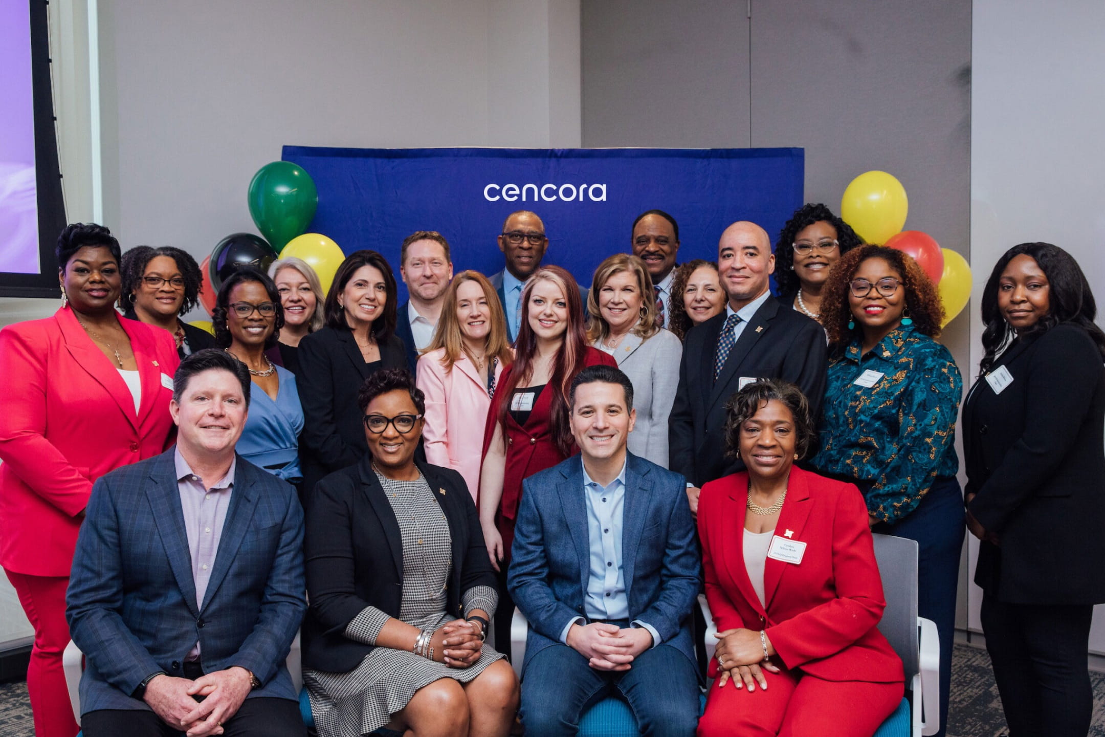 A diverse group of twenty people in an office posing for a photo in front of a dark blue Cencora banner with balloons on both sides. They are dressed in business attire and many wear name tags.