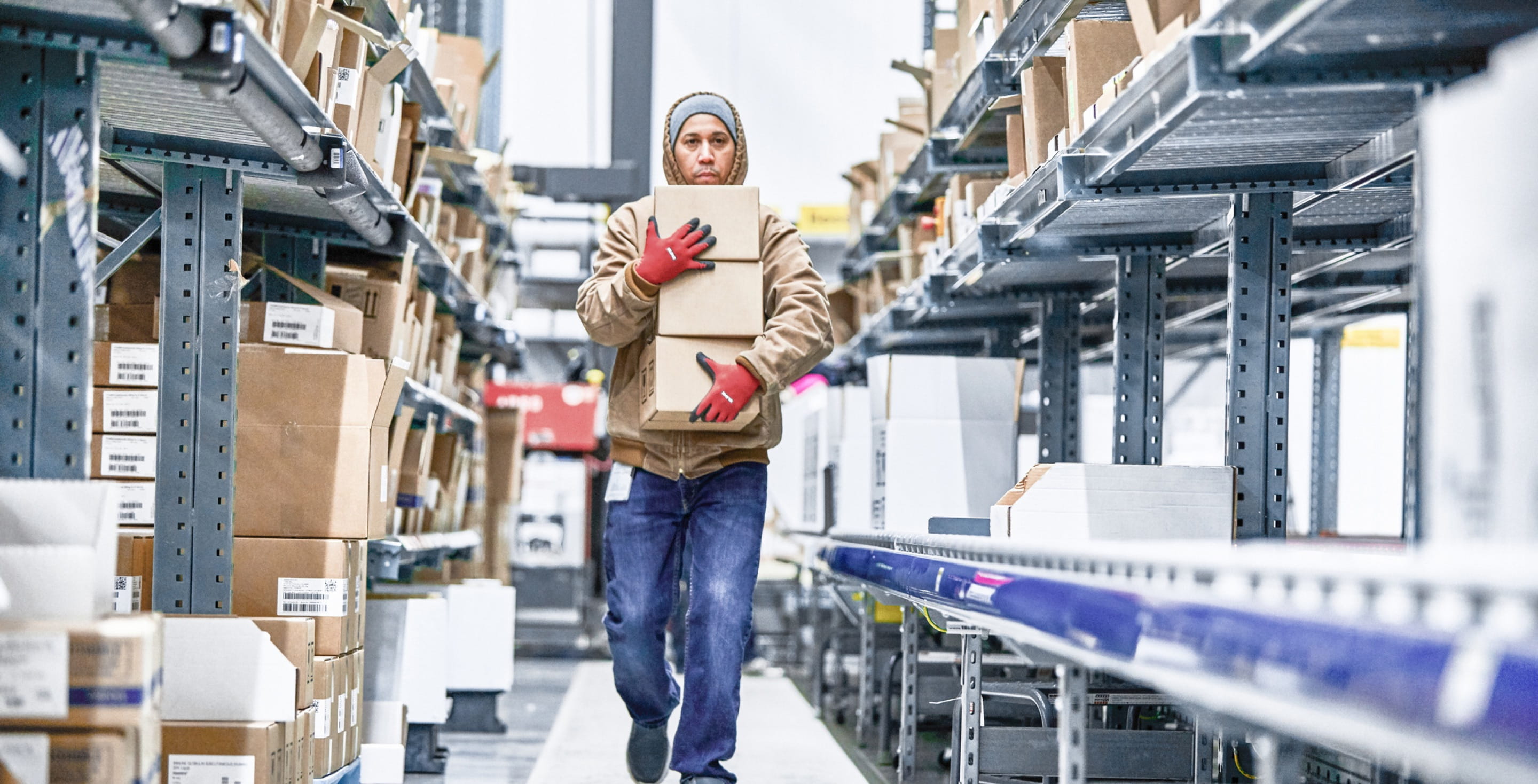 A warehouse worker wearing red gloves carrying three medium-sized boxes down an aisle. Boxes of various sizes line shelves on both sides of the worker.