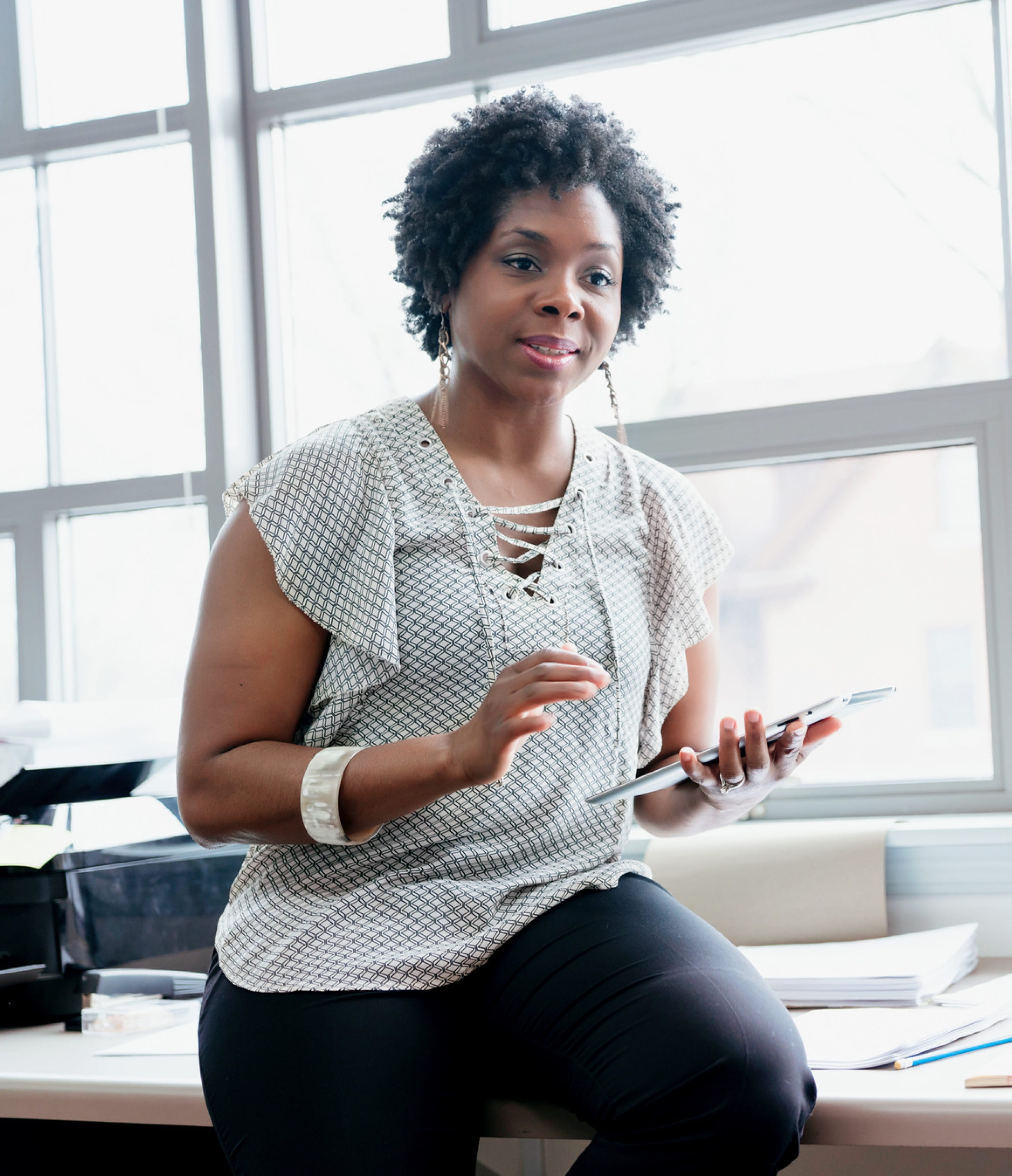 A dark-skinned person with short hair holding a tablet device leans on a desk while having a conversation with a person who is out of frame.