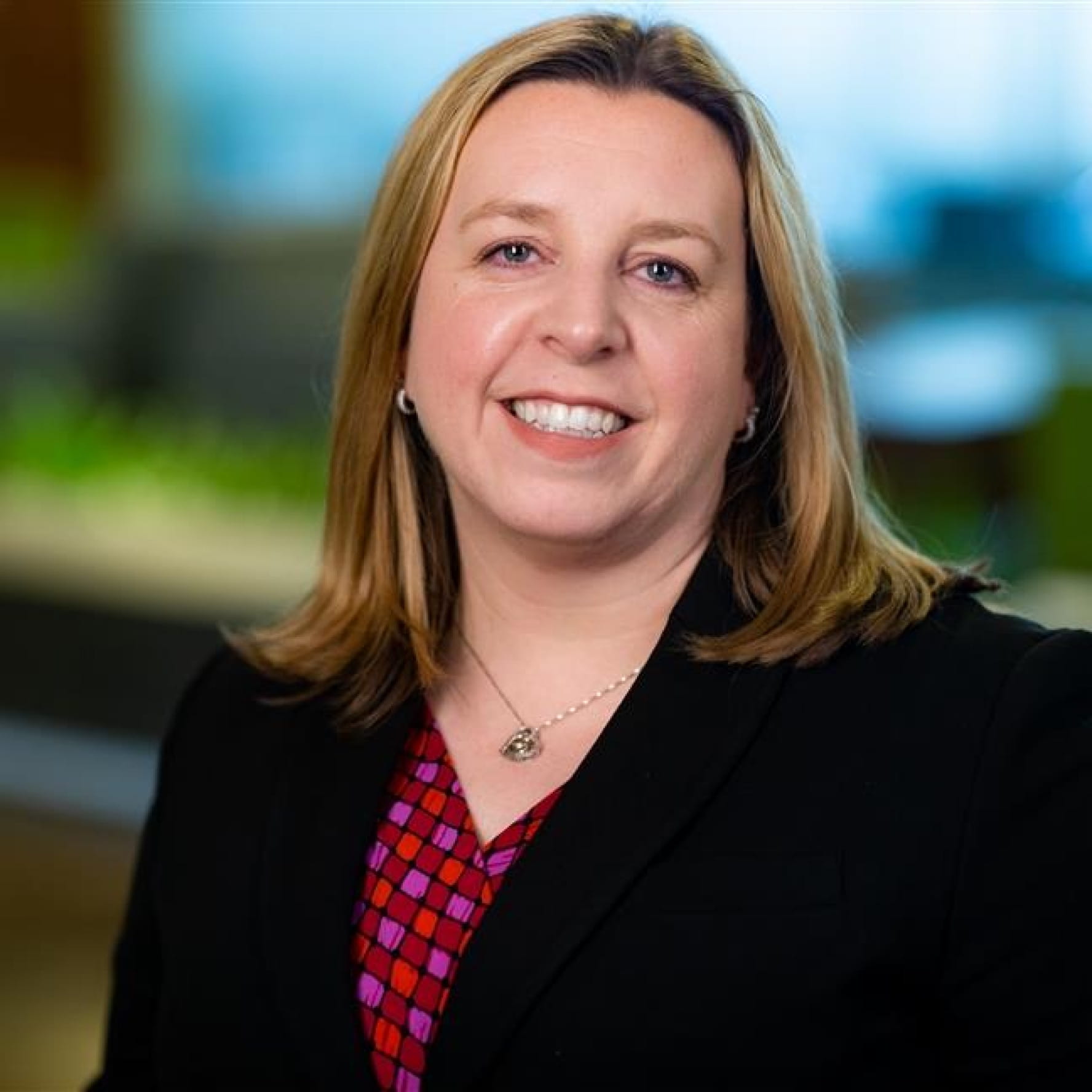 Photograph of Jennifer Dubas. Jennifer, a light-skinned person with blonde hair and blue eyes, is smiling at the camera. She wears hoop earrings, a pink shirt, and a black jacket. She is in front of a colorful background that is out of focus.