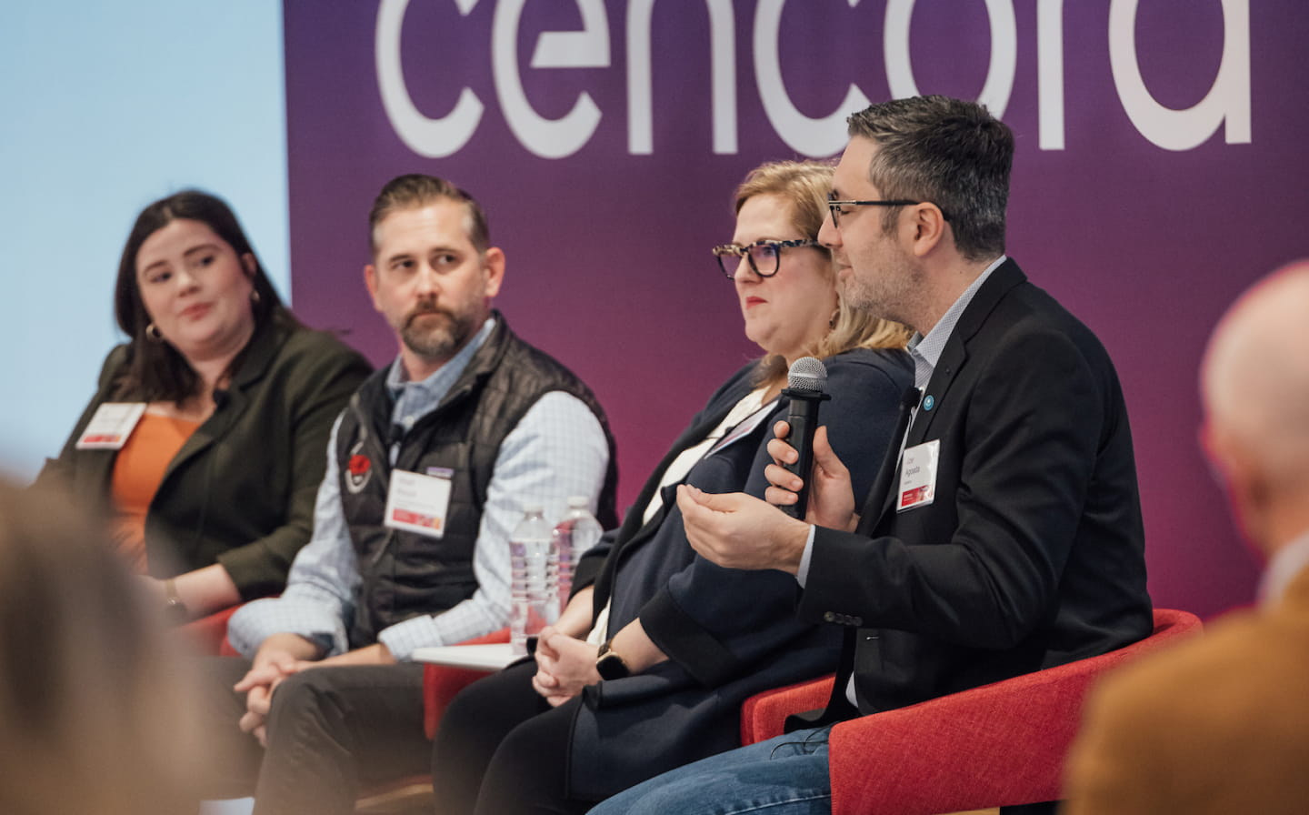 Four speakers with name tags sharing a stage during an event sponsored by Cencora. A person on the far right speaks into a microphone while two others on stage look at them.