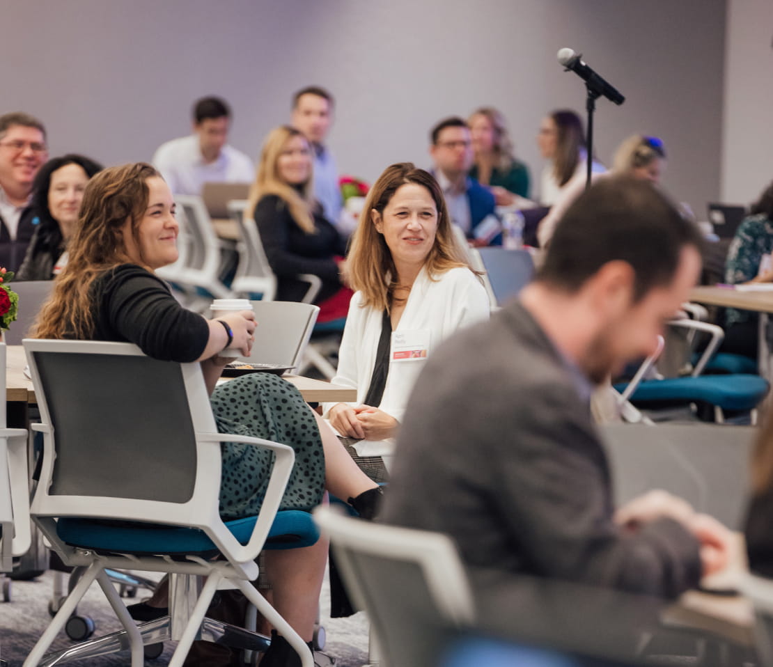 A crowd of people sitting in a conference room during a presentation. A microphone stands near the center of the audience as they wait for someone to speak.