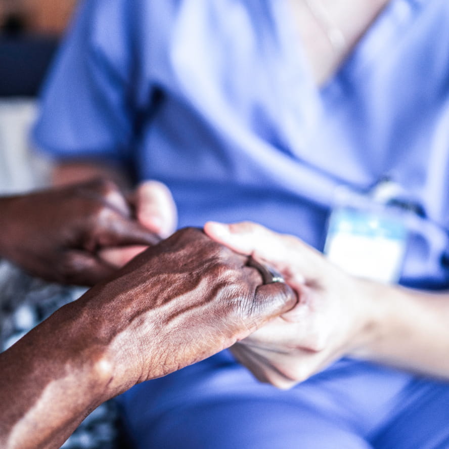 A dark-skinned person holding both hands with a light-skinned person wearing scrubs and a name tag. No faces are visible.