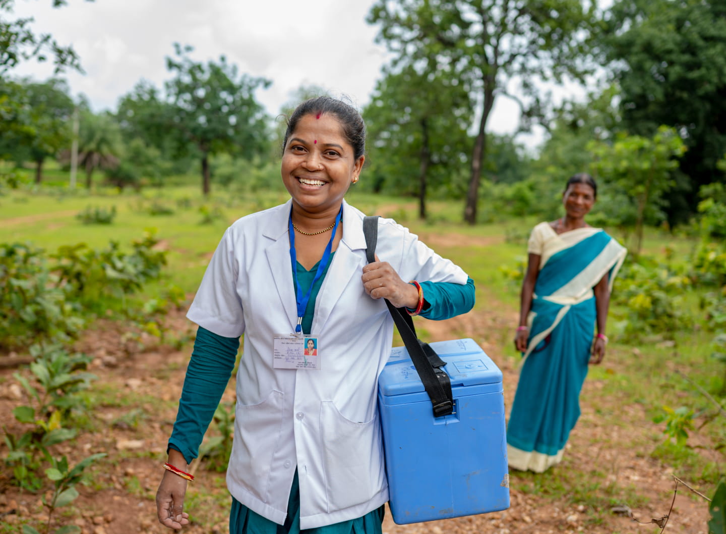 A dark-skinned medical professional smiling in an outdoor rural setting with lots of green vegetation holds a medical travel cooler. Behind them a dark-skinned person smiles and wears a green and white wrapped dress.