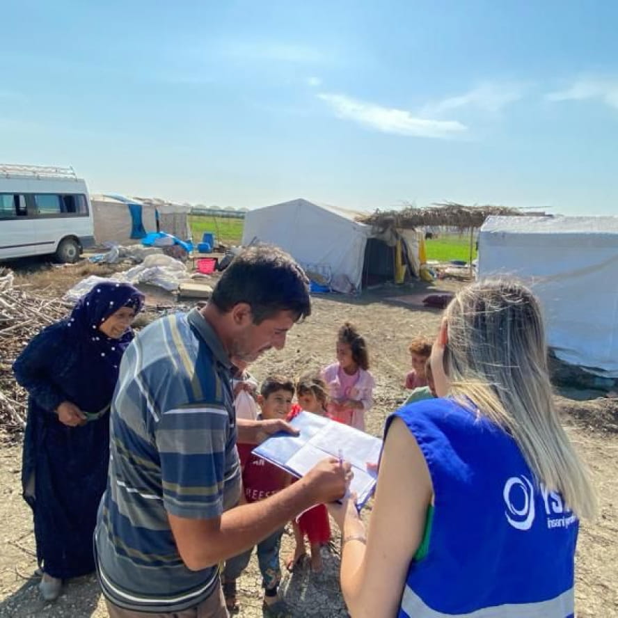 Two adults reviewing information on a clipboard while another adult watches over six children. Behind them is a white van, a pile of supplies, and two white humanitarian canopy shelters.