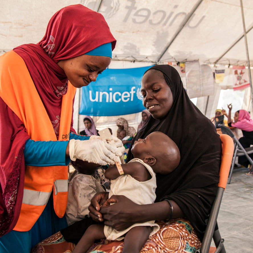 A dark-skinned person wearing an orange Unicef vest putting a wristband on a small dark-skinned child inside of a white canopy shelter. A third person wearing a black hijab holds the child.