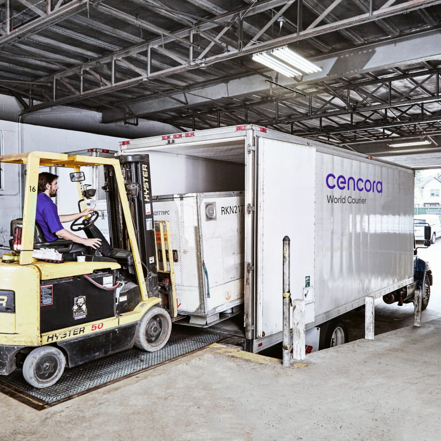 A light-skinned worker operating a forklift loading a thermal container into a truck that has the Cencora World Courier logo printed on the side.