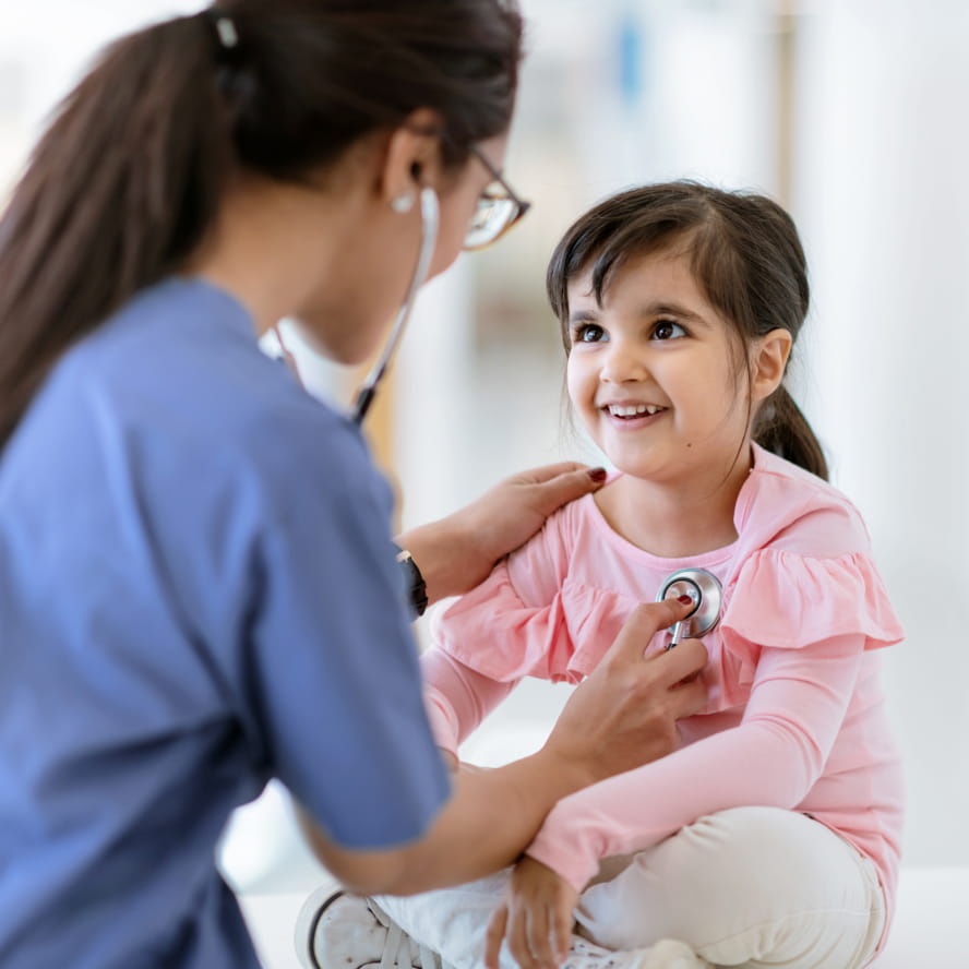 A medical professional wearing scrubs measuring a young child’s heartbeat with a stethoscope. The child wears a pink shirt and white pants and is smiling at the medical professional.