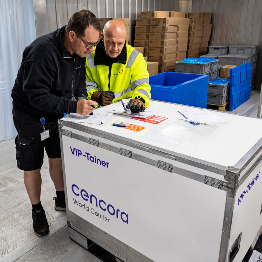 Two workers, one wearing a black fleece jacket and one wearing a yellow safety jacket, reviewing paperwork on a large white thermal container. It has the Cencora and World Courier logos printed on the side along with the words “VIP-Tainer”.
