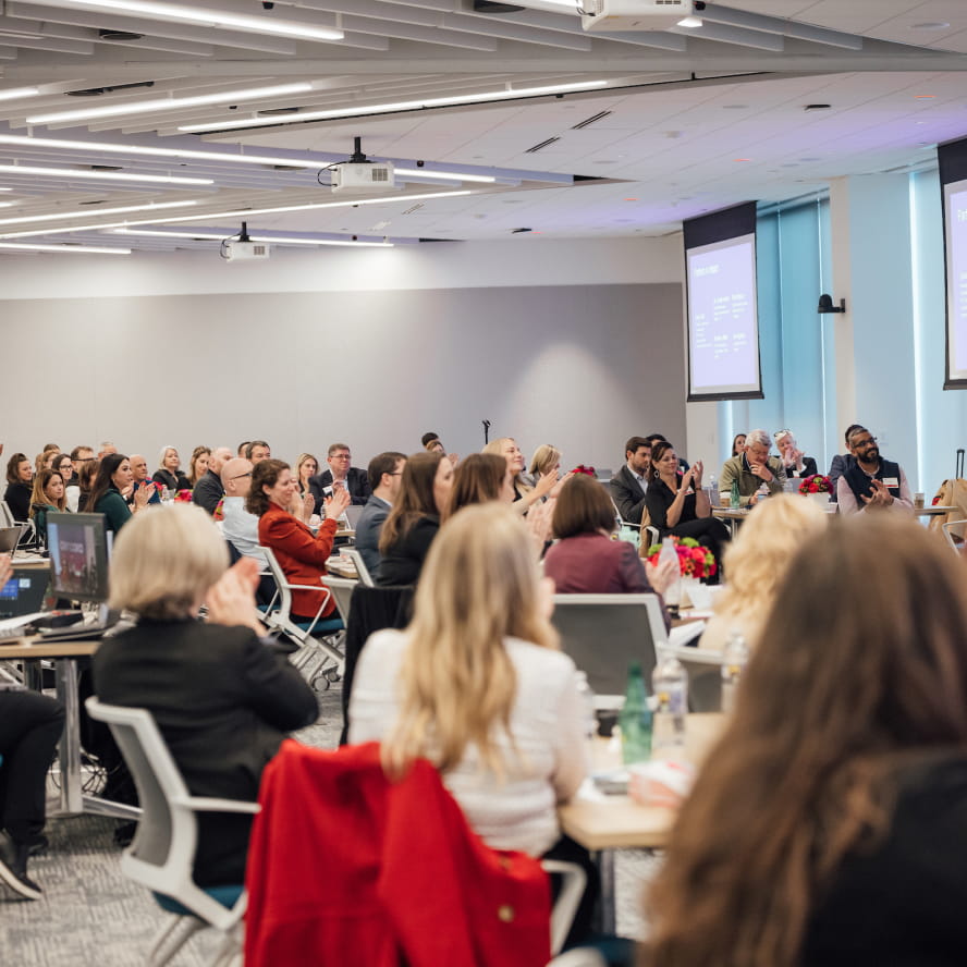 Dozens of professionals sitting in a large corporate conference room watch an event speaker who is out of frame.