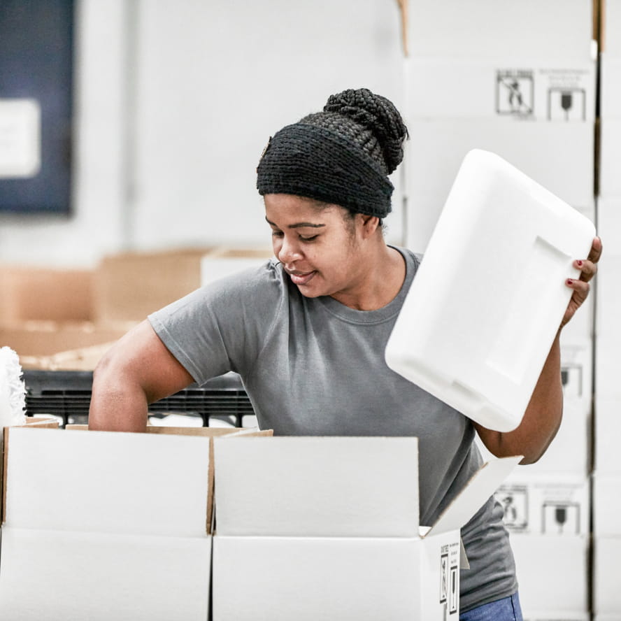 A dark-skinned worker in a warehouse packing pharmaceuticals into boxes. They wear a gray t-shirt and have their black braided  hair pulled up and wrapped over their head.