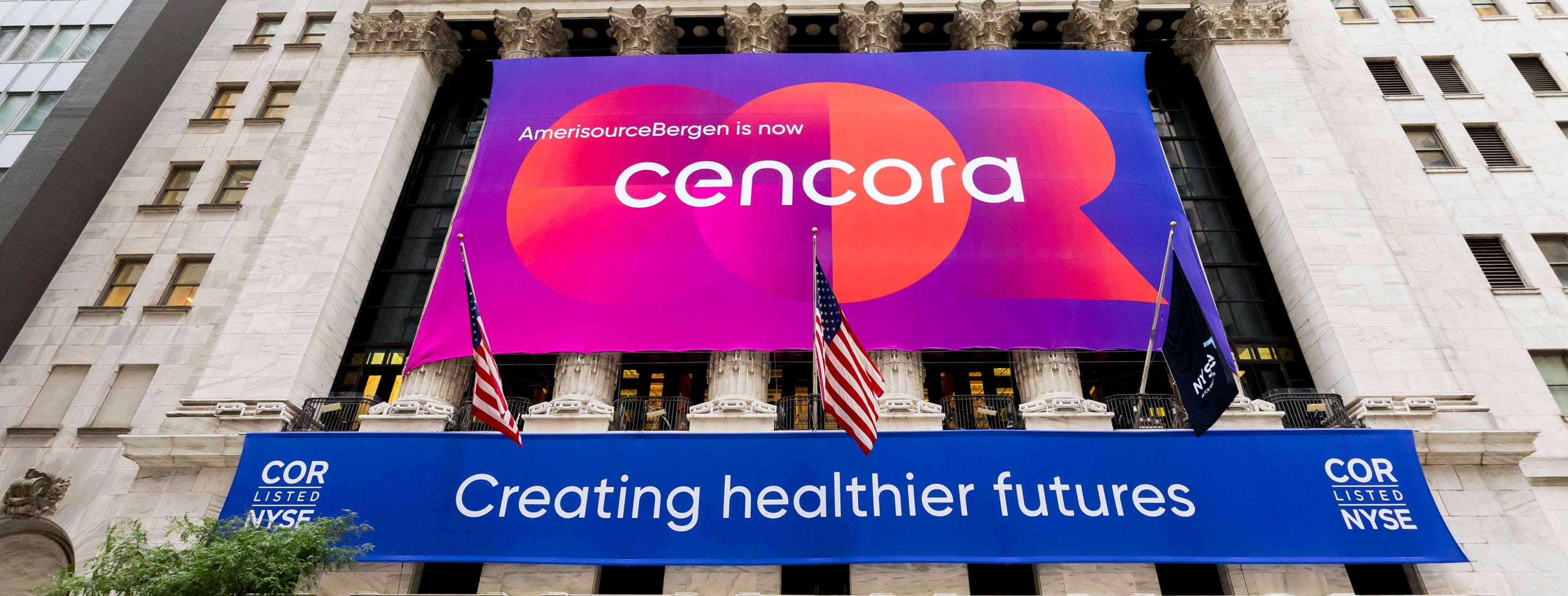 A Cencora banner hanging in front of the New York Stock Exchange building announces, “AmerisourceBergen is now Cencora.” A shorter banner below it reads, “Creating healthier futures.” It also lists Cencora’s ticker symbol: COR.