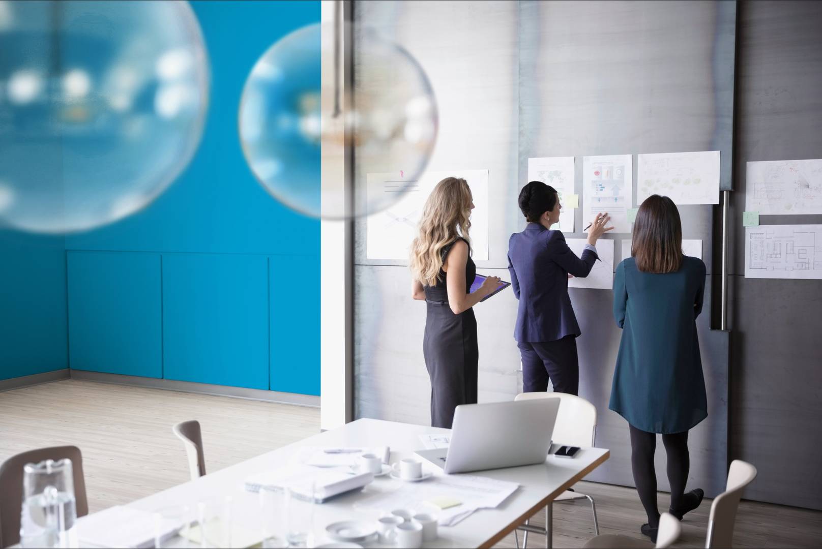 Three office workers dressed in business attire looking over some paper documents that are adhered to a conference room wall.