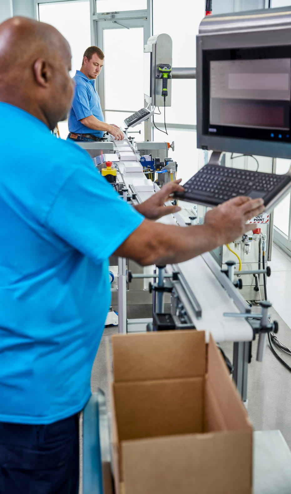 Two workers in blue shirts monitoring an automated pharmaceutical assembly line as small white boxes move down the line. They both stand in front of computers.