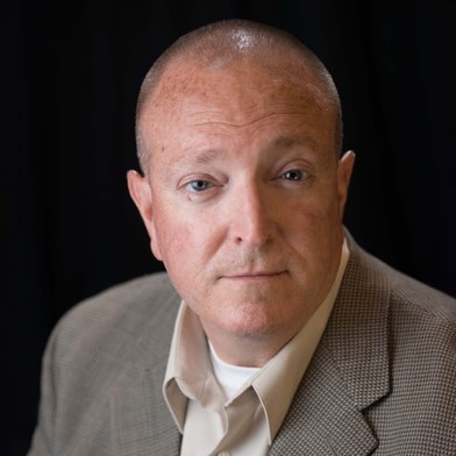Photograph of Robert Crow. Robert, a light-skinned person with very short brown hair and blue eyes, is looking into the camera. They wear a tan collared shirt beneath a brown textured suit jacket. They are in front of a black background.