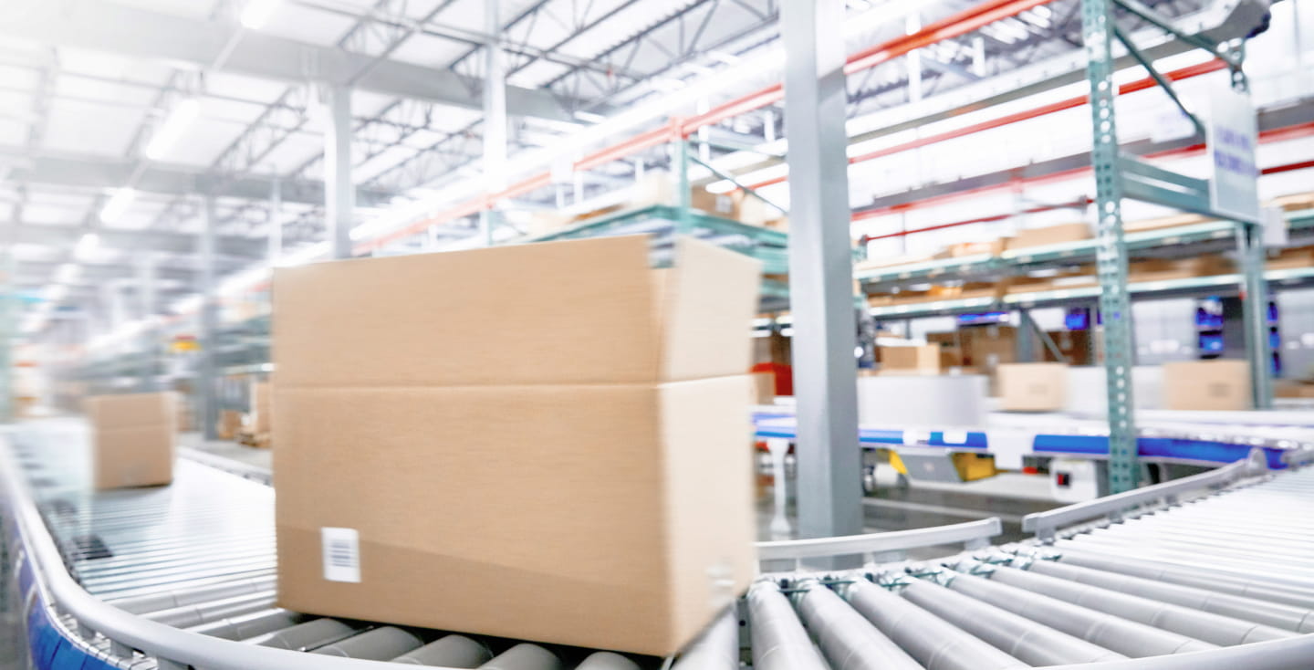 A large brown box moving down a conveyor belt inside of a large, bright warehouse factory.