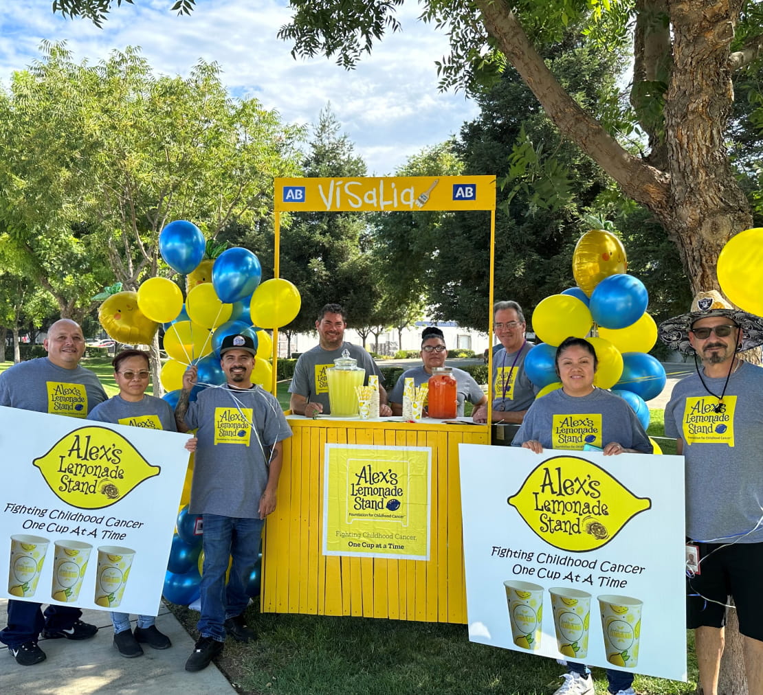 Eight Cencora volunteers posing outside around a yellow lemonade stand. They wear t-shirts and hold posters that read “Alex’s Lemonade Stand - Fighting Childhood Cancer One Cup At A Time"