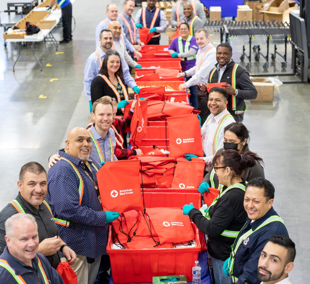 Twenty Cencora volunteers posing in a warehouse while packing emergency supplies into red canvas bags with the American Red Cross logo on them.
