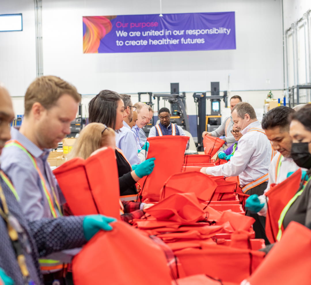 Cencora volunteerspacking emergency supplies into red canvas bags inside a warehouse. A banner on the wall behind them reads: Our Purpose. We are united in our responsibility to create healthier futures.
