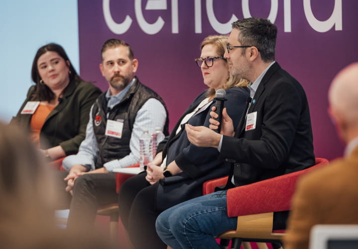 Four speakers with name tags sharing a stage during an event sponsored by Cencora. A person on the far right speaks into a microphone while two others on stage look at them.