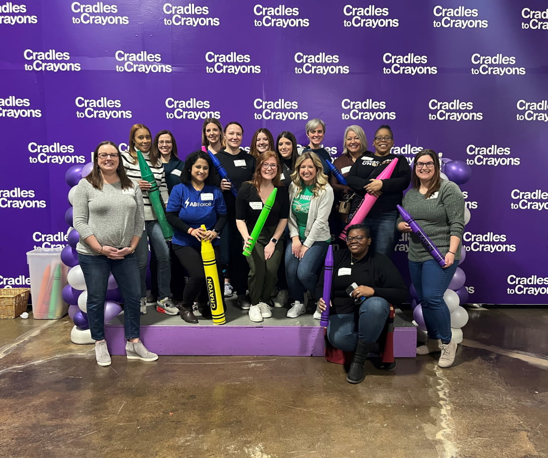 Fifteen volunteers posing while holding large colorful crayons in front of a purple wall. The “Cradles to Crayons” logo is in white letters and repeats across the wall behind them.