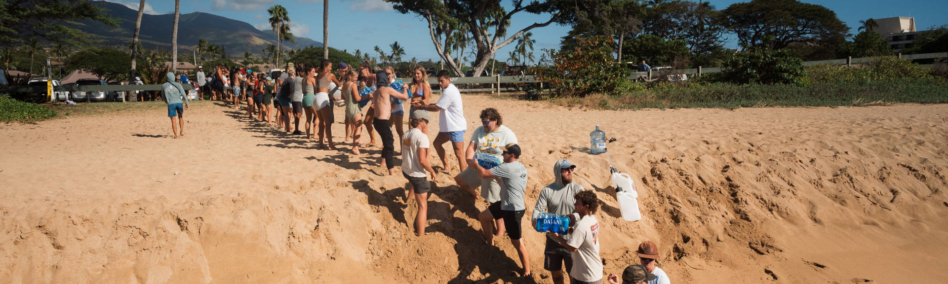 A large group of volunteers passing large packages of Dasani water bottles from one end of the row of people to the other. They are standing on a sandy and hilly beach in a tropical setting surrounded by palm trees.